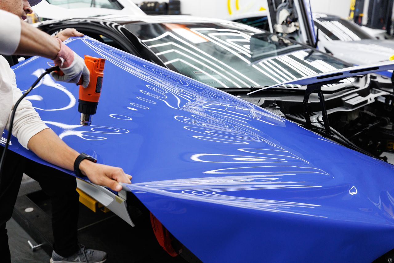 services-02 A technician uses a heat gun to carefully apply a blue vinyl wrap to a car in an auto workshop.