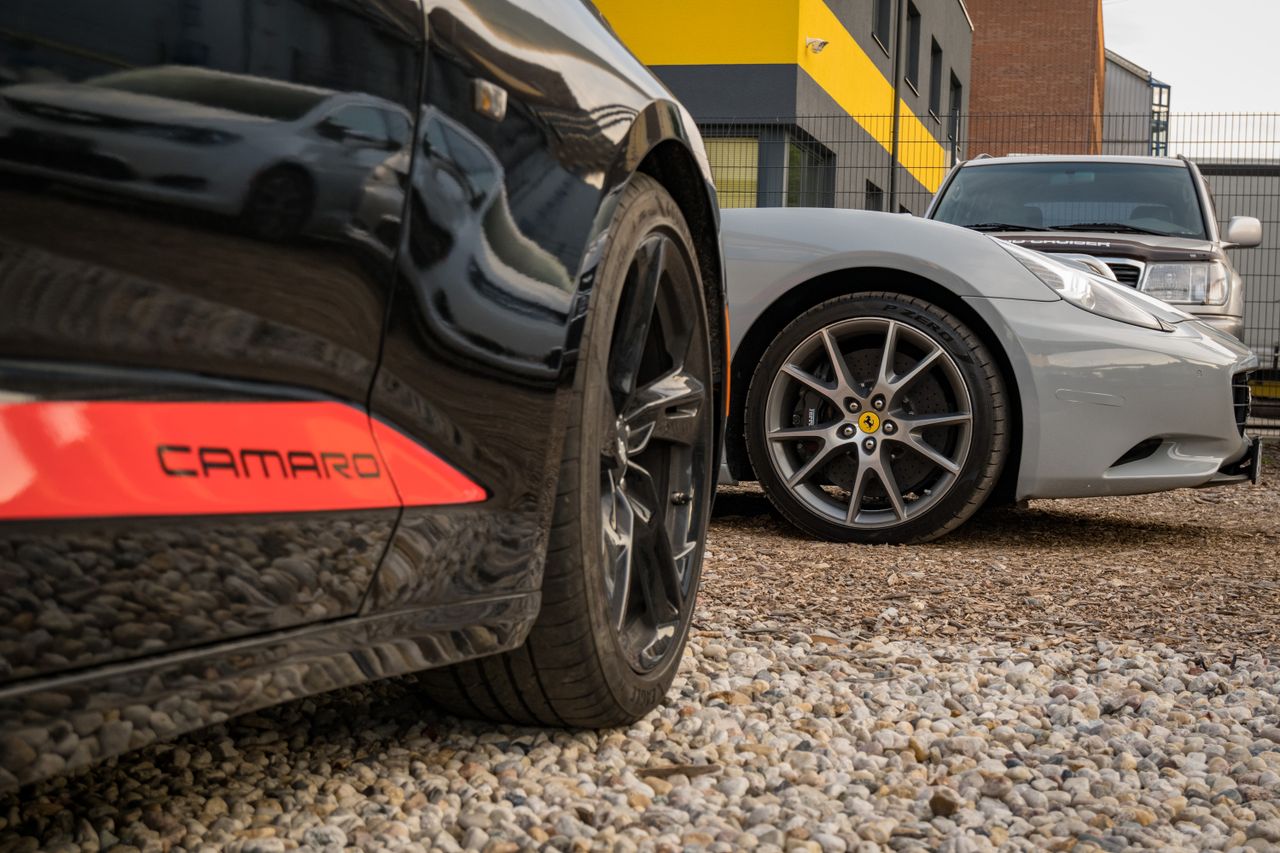 Close-up of luxury cars on gravel with industrial backdrop, featuring Camaro and sports car.