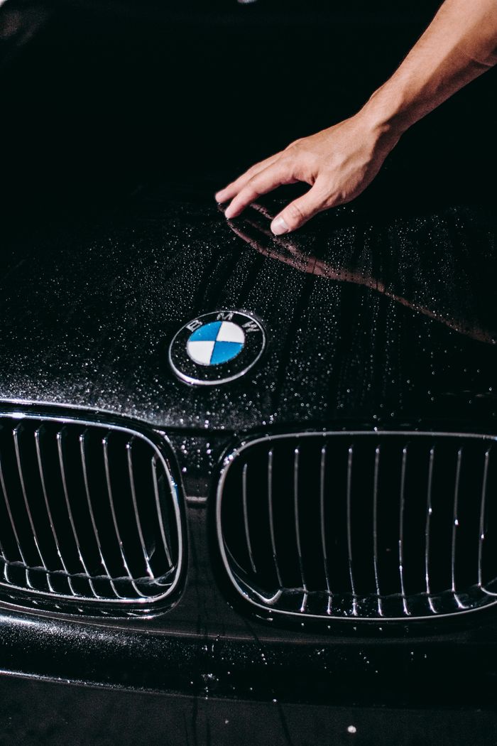 about-01 Close-up of a luxurious BMW car hood, showcasing the iconic emblem with water droplets and a hand.